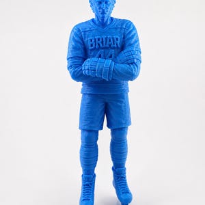 Op de afbeelding: Een blauw beeldje van een hockeyspeler in een trui met de tekst "BRIAR 44" erop. De figuur draagt een korte broek, lange sokken en schaatsen, met gekruiste armen. De figuur staat op een wit oppervlak.