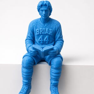 Op de afbeelding: Blauw beeld van een ijshockeyspeler die een boek leest. De speler draagt een trui met "BRIAR 44", ijshockeyschaatsen en kniebeschermers. Het beeld staat tegen een witte achtergrond.