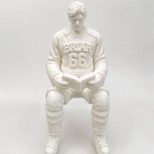 Op de afbeelding: Een wit 3D-geprint beeldje van een ijshockeyspeler die zit en een boek leest. De speler draagt een shirt met de tekst BRIAR en het nummer 66 op de borst, samen met een ijshockeybroek, sokken en schaatsen. Het gehele beeldje is monochroom.