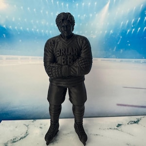 Op de afbeelding: Een matzwart 3D-geprint beeldje van een ijshockeyspeler die met gekruiste armen staat. De speler draagt een shirt met de tekst BRIAR en het nummer 66 op de borst, een ijshockeybroek en schaatsen. Het beeldje heeft een getextureerd oppervlak.