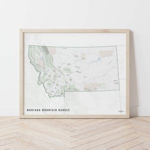 May include: A framed map print showing the mountain ranges of Montana. The map uses green, grey, and beige to display topographical details. The text "MONTANA MOUNTAIN RANGES" is printed in black at the bottom. The frame is a light wood colour.