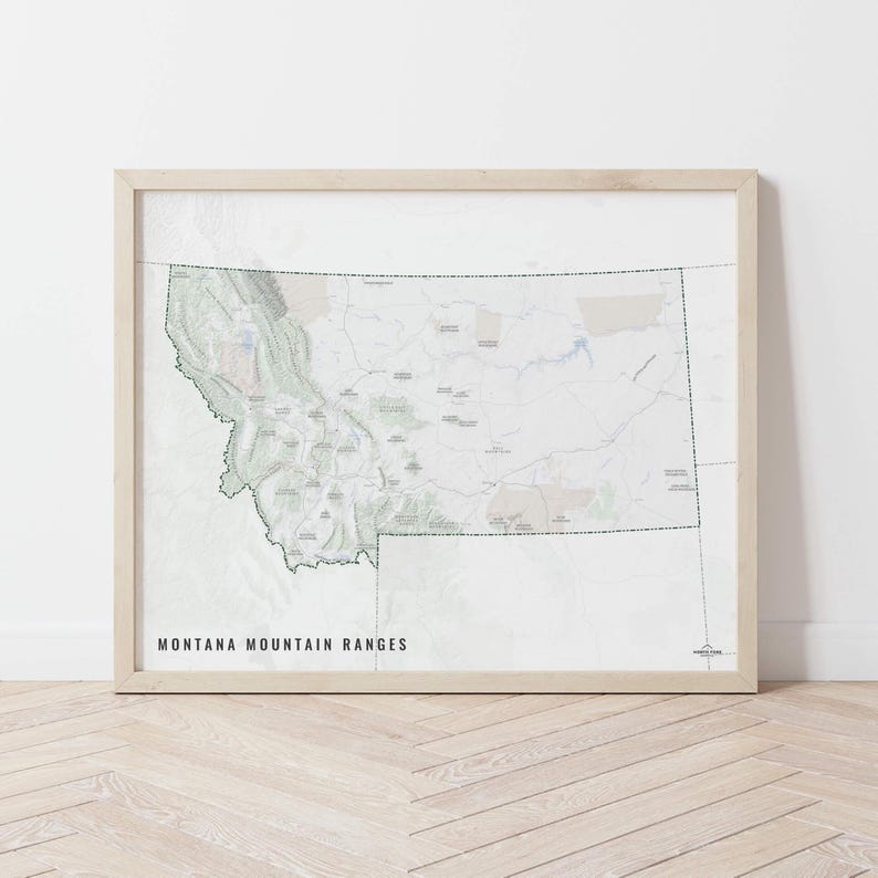 May include: A framed map print showing the mountain ranges of Montana. The map uses green, grey, and beige to display topographical details. The text "MONTANA MOUNTAIN RANGES" is printed in black at the bottom. The frame is a light wood colour.