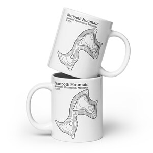 May include: Two white ceramic mugs with a black and white topographic map design of Beartooth Mountain in Montana. The text "Beartooth Mountain" and "Beartooth Mountains, Montana" is printed on the mug. The text "12,351 ft." is also printed on the mug.