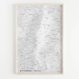May include: A framed topographic map print of the Bitterroot Valley in grayscale. The map features detailed contour lines and place names. The frame is a light wood color, and the text "BITTERROOT VALLEY" is printed in black at the bottom.