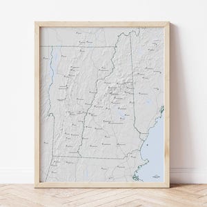 May include: A framed map of a state, featuring a detailed topographical representation. The map is predominantly white with green and blue accents, and is encased in a light-colored wooden frame. The map includes numerous place names and geographical features.