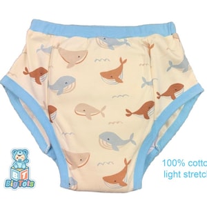 May include: Cream-colored baby training pants with a pattern of brown, gray, and beige whales and blue waves. The edges are trimmed with light blue. The text "100% cotton light stretch" is printed on the lower right.