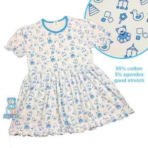 Puede incluir: Vestido blanco con ribete azul y un estampado de ositos de peluche, patos y otros juguetes azules. El vestido tiene mangas cortas y un dobladillo con volantes. El texto de la imagen dice "95% algodón 5% spandex buena elasticidad".