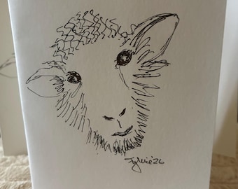 May include: A white greeting card featuring a black ink line drawing of a sheep head. The sketch shows the sheep with curly wool on its forehead, two ears, and dark eyes. The artist signature Sylvie26 is written in black ink below the sheep head.