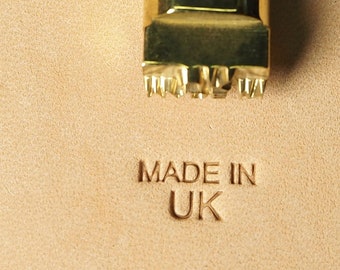 Könnte beinhalten: Nahaufnahme eines goldenen Stempels mit einem Sternmuster und den Worten "MADE IN UK", die in ein Stück braunes Leder geprägt sind.