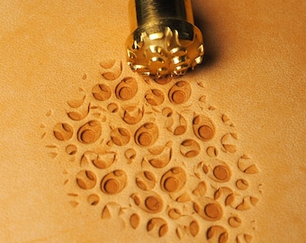 Può includere: Un primo piano di uno strumento per la stampa del cuoio con una testa in ottone e un design dettagliato. Lo strumento viene utilizzato per creare uno schema di cerchi e volute sul cuoio.