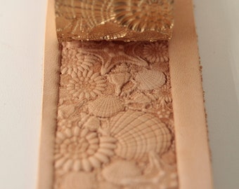 May include: A close-up of a leather strip with a seashell design embossed into the surface. A metal stamp with a matching design is shown above the leather.