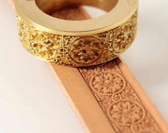 Könnte beinhalten: Ein goldener Stempel aus Metall mit einem detaillierten Blumenmuster. Der Stempel wird verwendet, um ein Muster auf ein Stück Leder zu erstellen.