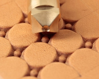 May include: Close-up of a leather crafting tool with a brass head, pressing into tan leather. The leather is embossed with a pattern of circles and small spheres, creating a textured surface. The tool is silver and brass.