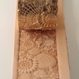 May include: A close-up of a leather strip with a seashell design embossed into the surface. A metal stamp with a matching design is shown above the leather.