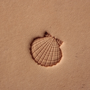 May include: A detailed, embossed seashell design on a tan leather surface. The shell is intricately carved with radiating lines and a textured appearance, creating a three-dimensional effect. The background is a plain, textured leather.