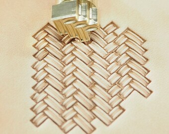 May include: A close-up of a leather stamping tool being used to create a woven pattern on a piece of leather. The tool is made of brass and has a square head with a raised design.