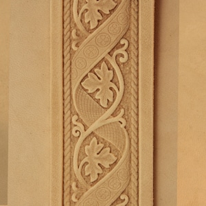 May include: A close-up of a light brown leather strip with a decorative pattern of leaves and vines. The pattern is embossed into the leather and has a raised texture.