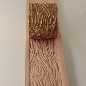 Puede incluir: Un sello de metal dorado con un patrón de veta de madera se utiliza para crear una textura de veta de madera en un trozo de cuero.