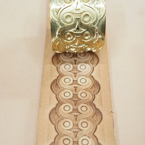 May include: A brass leather stamping tool with a decorative pattern is being used to create a design on a piece of leather.