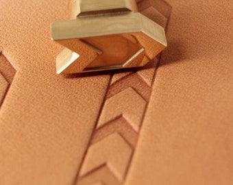 Può includere: Uno strumento di stampaggio in pelle color oro con un motivo a chevron. Lo strumento è appoggiato su un pezzo di pelle marrone.