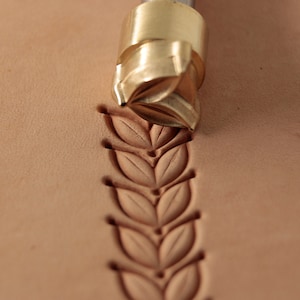 Pode incluir: Uma ferramenta de carimbo de couro de latão com um design de folha. A ferramenta é usada para criar um padrão decorativo em um pedaço de couro.