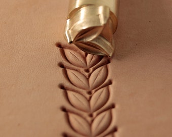 May include: A close-up of a brass leather stamping tool with a leaf design. The tool is being used to create a decorative pattern on a piece of leather.