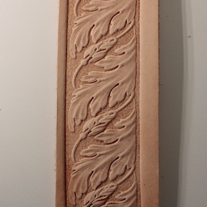 May include: A close-up of a light brown leather strip featuring an embossed leaf design. The leaves are arranged vertically, with a textured background. The edges of the leather are smooth and a lighter shade of brown.