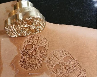 Op de afbeelding: Een messing stempel met een gedetailleerd schedelontwerp, 30 mm bij 20 mm groot. De stempel wordt gebruikt om indrukken op leer te maken. De afbeelding toont twee indrukken van de stempel op een stuk leer.