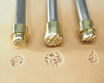 May include: Three metal leather stamping tools with brass heads. The tools are arranged horizontally on a light brown leather surface. The heads of the tools have different floral designs.