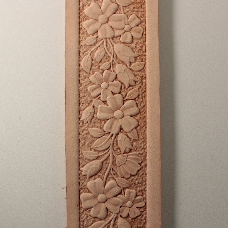 May include: A close-up of a leather strip with a carved floral design. The design features a repeating pattern of flowers and leaves, all carved in relief. The leather is a light brown color.