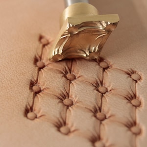 Può includere: Uno strumento di stampaggio in ottone per cuoio con un motivo floreale viene utilizzato per creare un motivo decorativo su un pezzo di cuoio. Lo strumento viene premuto sul cuoio per creare una serie di punti in rilievo.