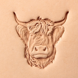 May include: A close-up of a leather stamp featuring a detailed carving of a Highland cow's head. The cow has a long, shaggy mane and a large, prominent nose.