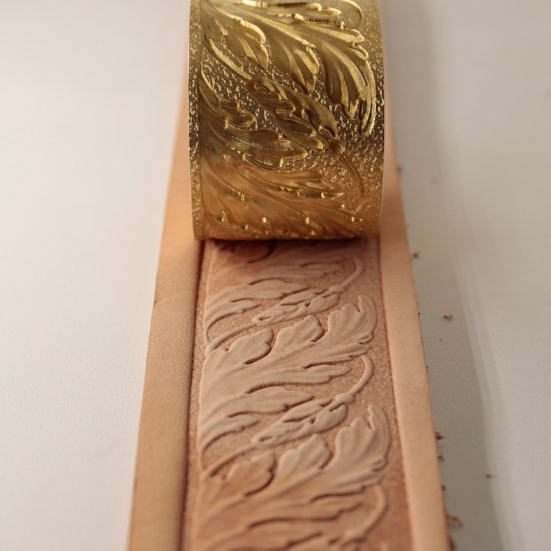 May include: A gold-coloured leather embossing tool rolling over a light brown leather strip. The tool has a detailed leaf pattern. The leather strip shows an embossed leaf design, created by the tool. The leather strip is approximately 2.5 cm wide.