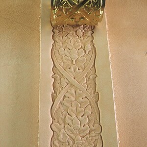 May include: Close-up of a tan leather strip with an embossed floral design. The design features leaves and intersecting lines, with a gold-colored metal piece at the top. The leather has a textured surface.