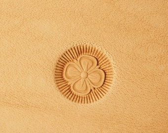 Può includere: Primo piano di una superficie in pelle marrone chiaro con un motivo floreale decorativo in rilievo al centro. Il fiore ha cinque petali e un piccolo cerchio al centro.