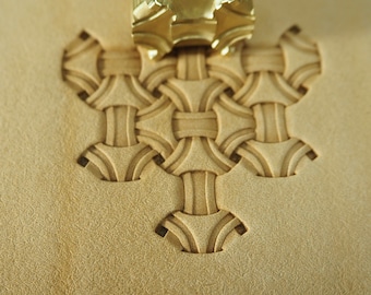 May include: A close-up of a leather stamping tool being used to create a woven pattern on a piece of leather. The tool is made of brass and has a detailed design.