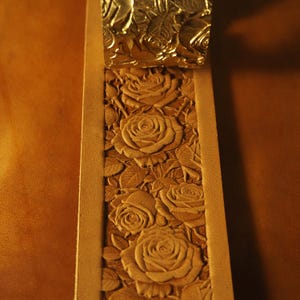 May include: A tan leather strip featuring a deeply embossed floral pattern of roses and leaves. A metallic gold-toned stamping tool with a matching rose design is positioned at the top of the leather, showing the process of creating the relief.