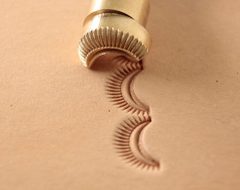 May include: A close-up of a brass leather stamping tool with a decorative design. The tool is being used to create a pattern on a piece of leather.