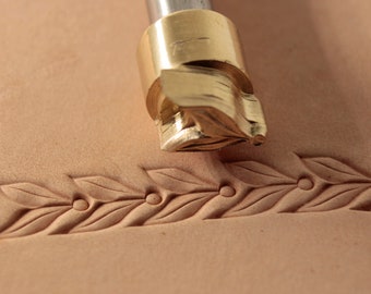 May include: A close-up of a brass leather stamping tool with a leaf design. The tool is being used to create a decorative border on a piece of leather.