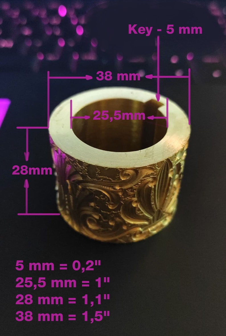 May include: A gold-coloured, cylindrical object with ornate floral and leaf patterns. The object has a key slot and measures 38 mm in diameter, 28 mm in height, and 25.5 mm in internal diameter. The key slot is 5 mm.