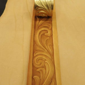 May include: Close-up of a handcrafted leather belt with intricate floral carving. The belt is a warm tan color with a detailed embossed design. The buckle is gold-colored and also features a floral pattern.