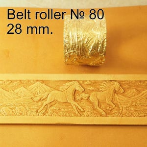 Könnte beinhalten: Ein goldfarbener Gürtelroller aus Metall mit der Aufschrift Belt roller № 80 28 mm. Der Roller zeigt ein detailliertes Prägemuster von zwei Pferden, die durch eine Berglandschaft laufen, welches auch auf einem beigen Lederstreifen zu sehen ist.