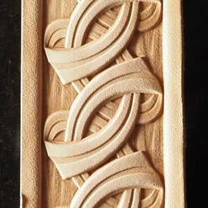 May include: A close-up of a leather strip with a detailed, intricate design of interwoven, curved lines. The design is carved into the leather, creating a raised, textured effect. The leather is a light brown color.