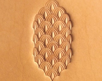 May include: A close-up of a leather stamping tool with a design of overlapping, rounded shapes resembling scales. The tool is made of metal and has a brown, textured finish.