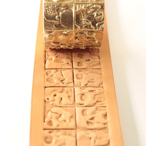 Pode incluir: Um carimbo de metal dourado com um desenho de caveira é pressionado sobre uma tira de couro castanho claro. O desenho do carimbo é replicado no couro, criando um padrão repetitivo de caveiras. A tira de couro está em um fundo branco.