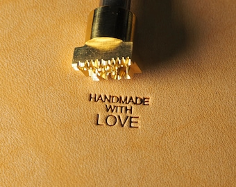 Può includere: Un timbro in metallo dorato con le parole "HANDMADE WITH LOVE" impresse su un pezzo di pelle marrone.