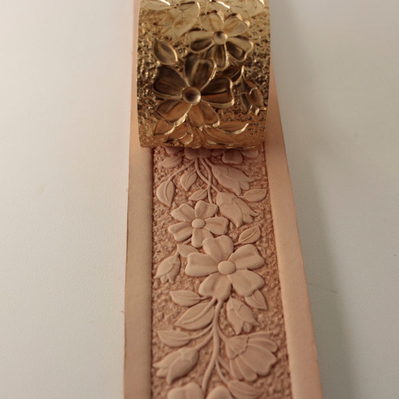 May include: A close-up of a leather strip with a floral design embossed into it. A metal stamp with a similar design is shown above the leather strip.