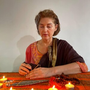 May include: A woman wearing a pink and brown shawl sits before a table with lit candles and an incense burner. A small silver elephant statue and prayer beads are also on the table. The scene suggests meditation and spirituality.