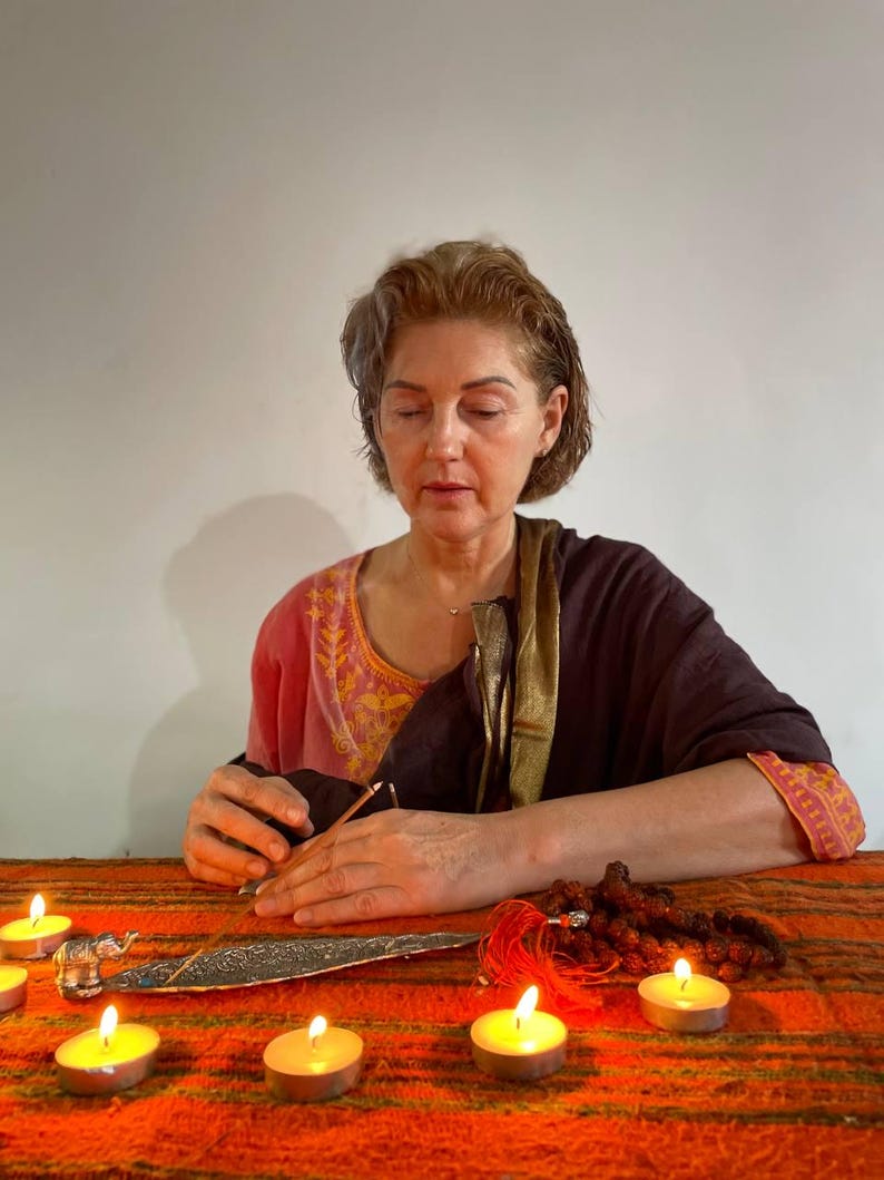 May include: A woman wearing a pink and brown shawl sits before a table with lit candles and an incense burner. A small silver elephant statue and prayer beads are also on the table. The scene suggests meditation and spirituality.