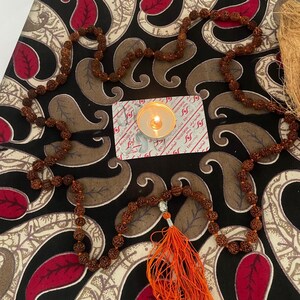 May include: A brown beaded mala necklace with an orange tassel, set on a patterned surface with red and brown paisley designs. A lit candle and a small card are also visible.